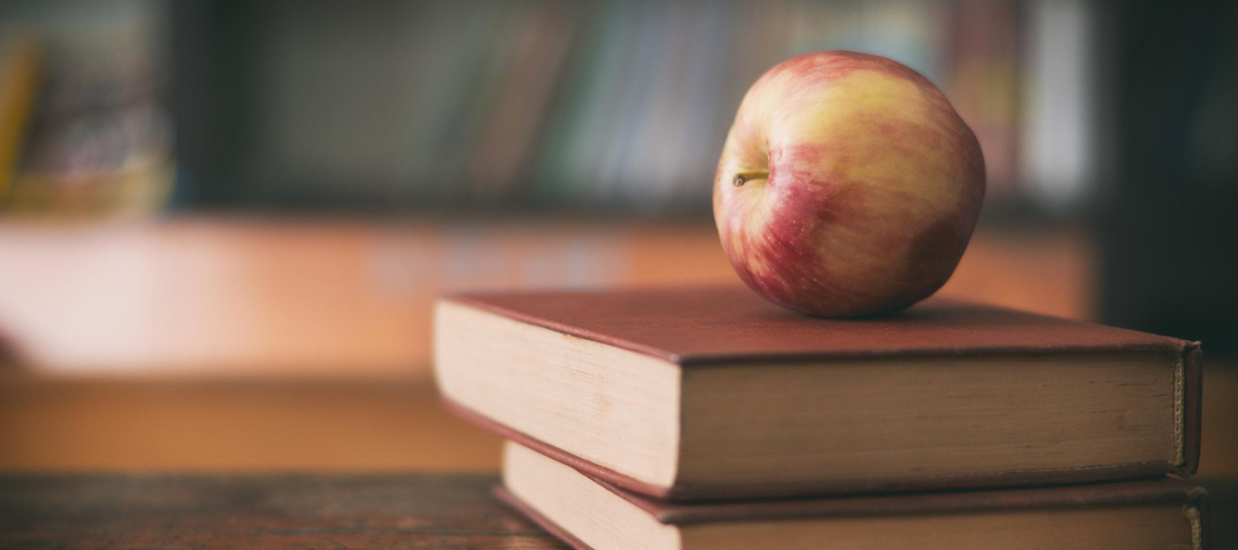 Apple sitting on a pile of books on a classroom desk.